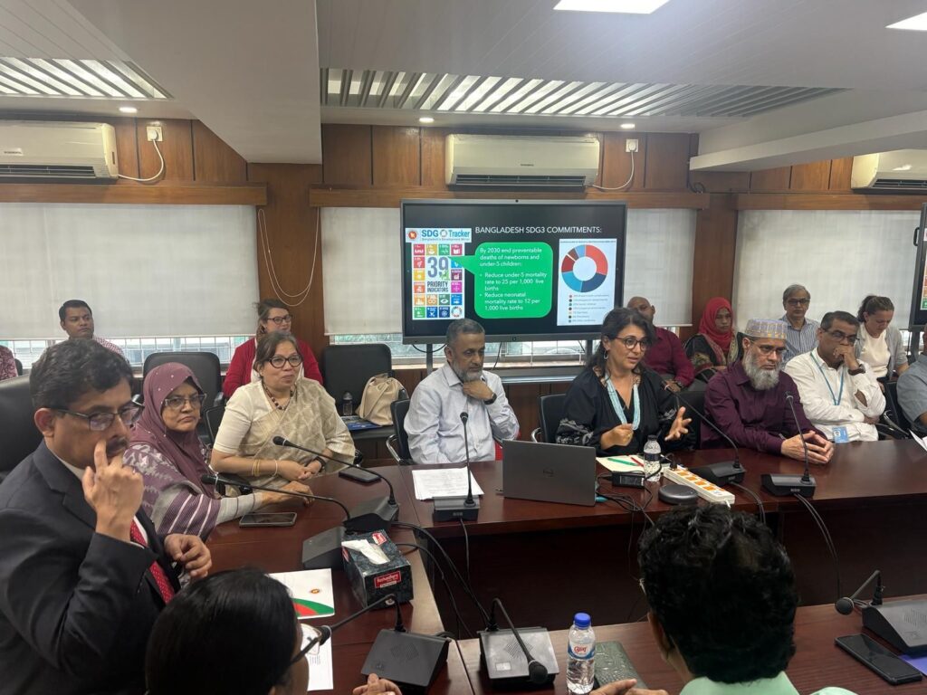 Large conference room with microphones on tables filled with people and a presentation screen in the background.